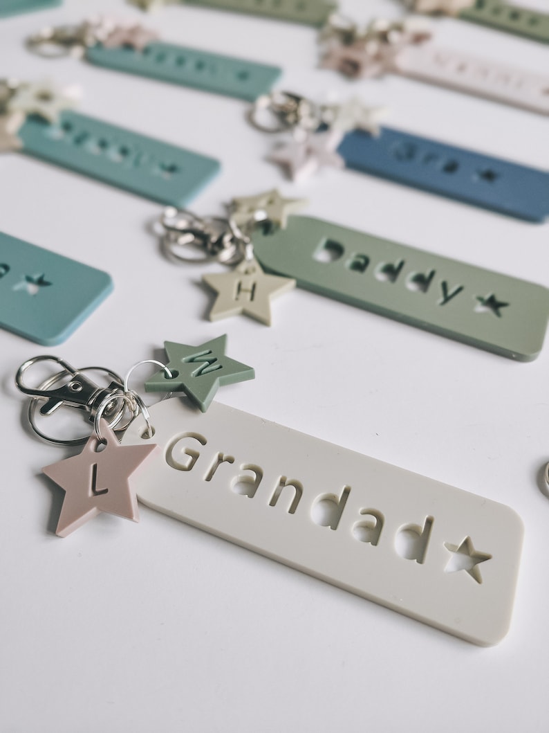 May include: A set of white, green, and blue keychains with star-shaped charms. Each keychain has a different name on it, including "Daddy", "Grandad", and "Bro".