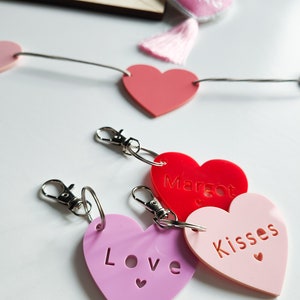 以下が含まれることがあります： 「Love」、「Margot」、「Kisses」と書かれたピンクと赤のハート型のキーホルダー3つ。キーホルダーは銀色のキーリングにぶら下がっています。