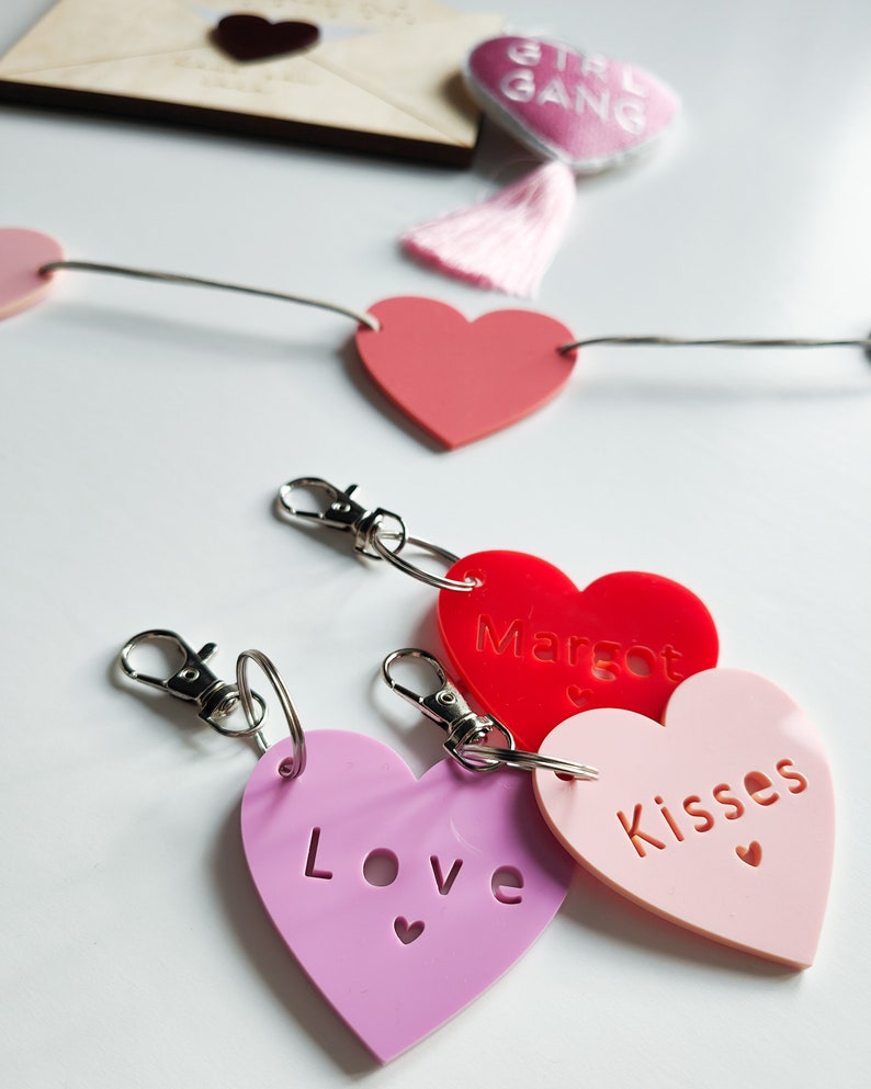 以下が含まれることがあります： 「Love」、「Margot」、「Kisses」と書かれたピンクと赤のハート型のキーホルダー3つ。キーホルダーは銀色のキーリングにぶら下がっています。