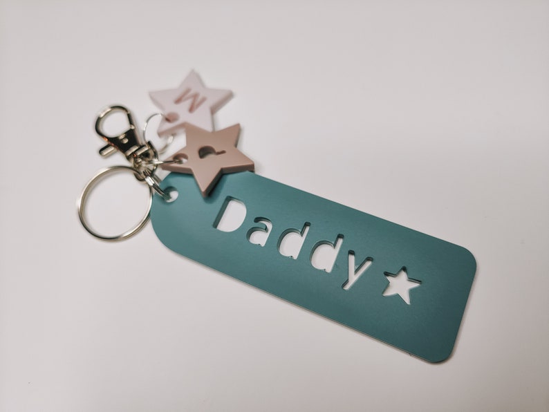 May include: Teal blue keychain with the word "Daddy" cut out and a small star. The keychain has two star-shaped charms attached, one pink and one beige.
