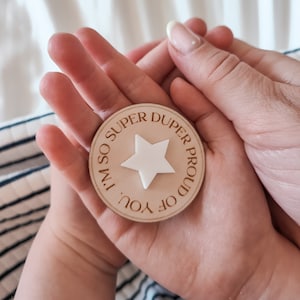 May include: A wooden circle with the text "I'm so super duper proud of you" and a white star in the centre. The circle is held in the hands of two people.