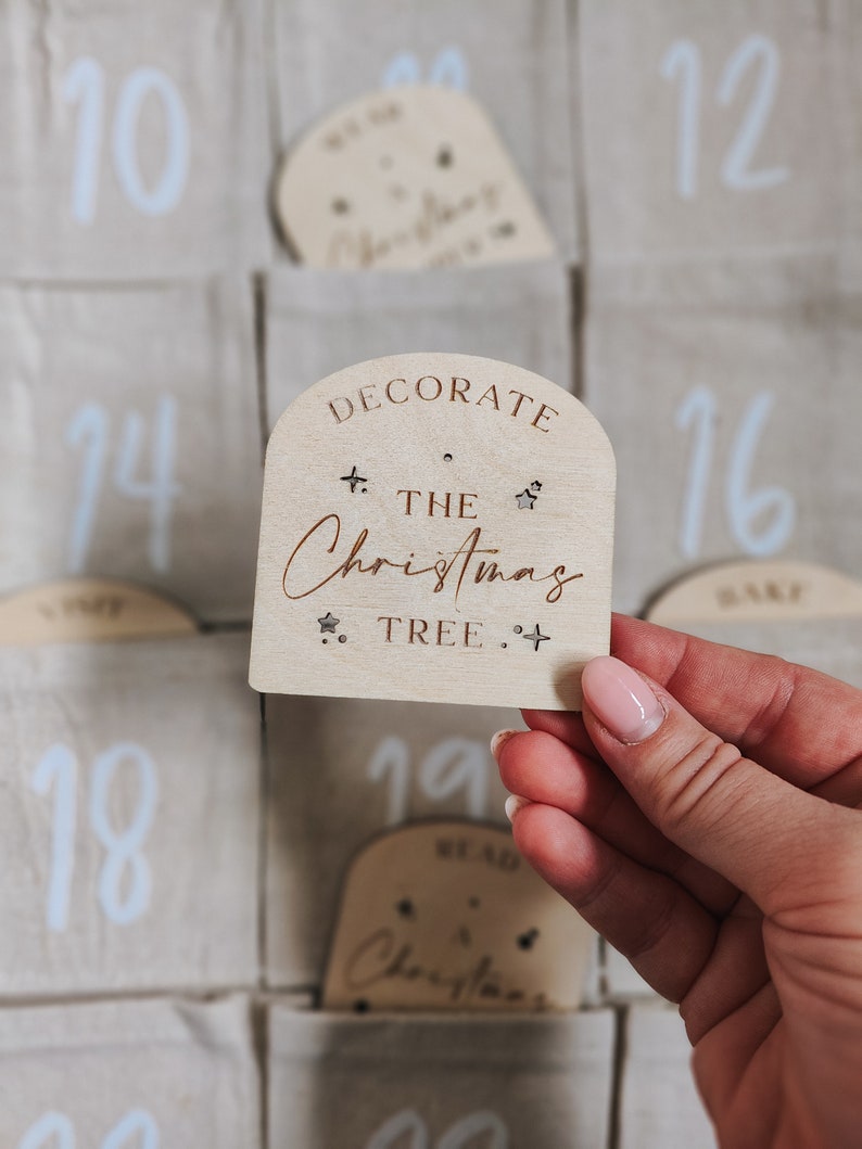 K&ouml;nnte beinhalten: Ein Adventskalender aus Holz mit hellbraunem Hintergrund. Der Kalender hat 24 nummerierte F&auml;cher, jedes mit einer kleinen Holzt&uuml;r. Die T&uuml;r in der Mitte ist ge&ouml;ffnet und zeigt eine kleine Holzplakette mit dem Text "Schm&uuml;cke den Weihnachtsbaum".