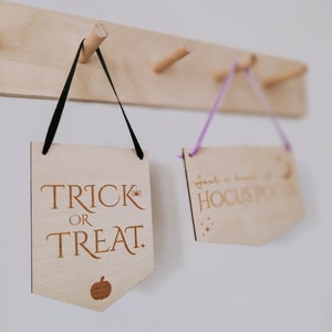 May include: Two wooden signs hanging on a wooden rack. The sign on the left says "Trick or Treat" with a small pumpkin icon. The sign on the right says "Just a little bit of Hocus Pocus" with stars.