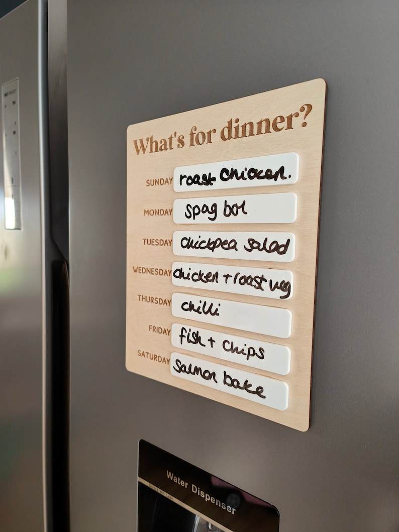 May include: A wooden sign with the text "What's for dinner?" and a list of meals for each day of the week. The meals are written on white labels and include roast chicken, spag bol, chickpea salad, chicken roast veg, chilli, fish and chips, and salmon bake.