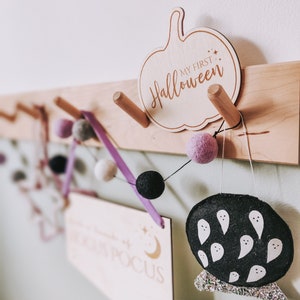 May include: Wooden wall decor with a pumpkin-shaped sign that says "My First Halloween". A purple ribbon with white and black pom-poms hangs from the sign. There is also a wooden sign that says "Hocus Pocus" and a black felt ghost with white ghost faces.