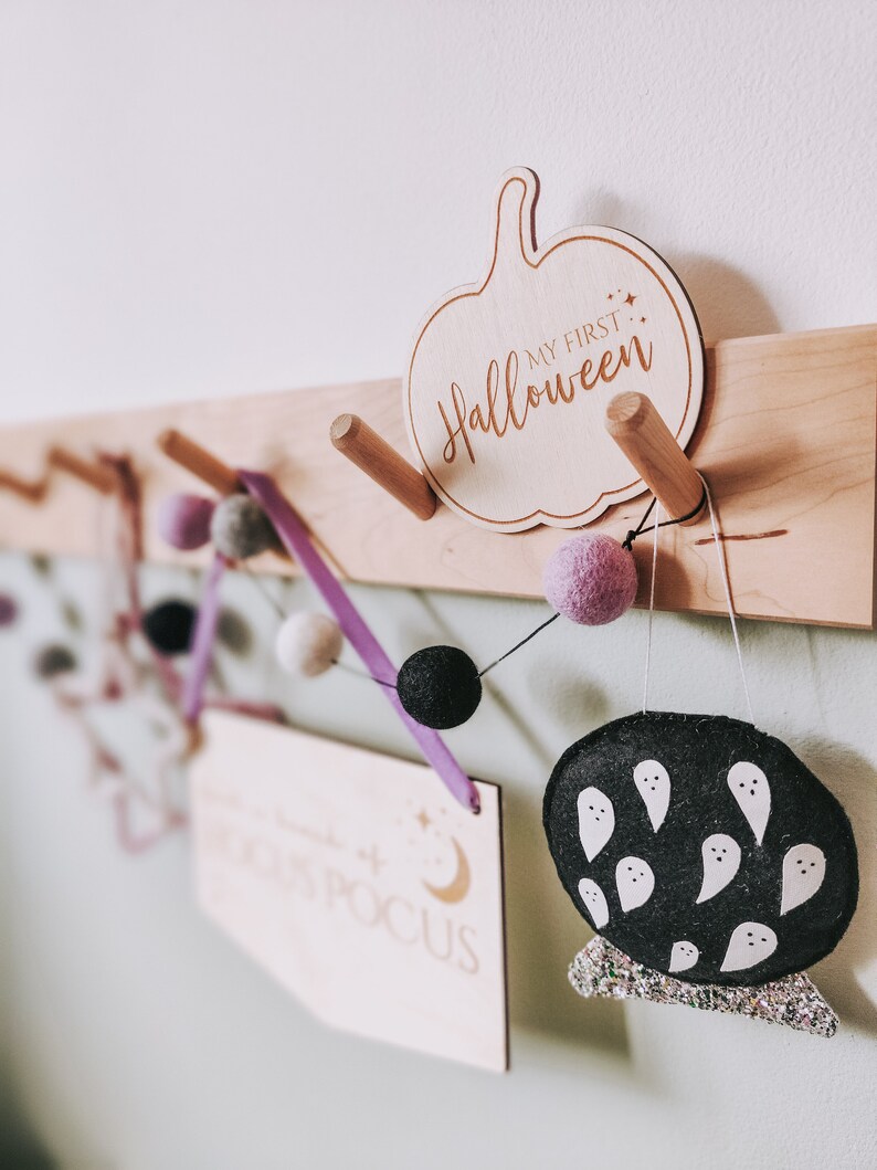May include: Wooden wall decor with a pumpkin-shaped sign that says "My First Halloween". A purple ribbon with white and black pom-poms hangs from the sign. There is also a wooden sign that says "Hocus Pocus" and a black felt ghost with white ghost faces.