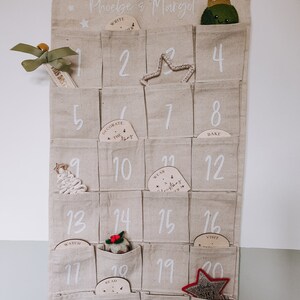 K&ouml;nnte beinhalten: Ein beigefarbener Stoff-Adventskalender mit 24 Taschen, jede mit einer Nummer und einem kleinen Holzschmuck. Die Ornamente haben verschiedene weihnachtliche W&ouml;rter darauf geschrieben, wie "Schreibe einen Brief", "Schm&uuml;cke den Baum", "Trage eine Weihnachtsm&uuml;tze" und "Besuche den Weihnachtsmann". Der Kalender ist mit Sternen verziert und die Namen "Phoebe & Margot" stehen oben drauf.