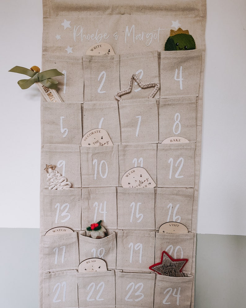 K&ouml;nnte beinhalten: Ein beigefarbener Stoff-Adventskalender mit 24 Taschen, jede mit einer Nummer und einem kleinen Holzschmuck. Die Ornamente haben verschiedene weihnachtliche W&ouml;rter darauf geschrieben, wie "Schreibe einen Brief", "Schm&uuml;cke den Baum", "Trage eine Weihnachtsm&uuml;tze" und "Besuche den Weihnachtsmann". Der Kalender ist mit Sternen verziert und die Namen "Phoebe & Margot" stehen oben drauf.