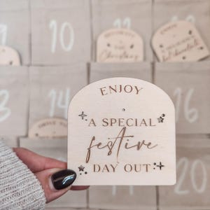 K&ouml;nnte beinhalten: Ein Adventskalender aus Holz mit einem Ausschnitt f&uuml;r einen festlichen Tag. Der Kalender hat einen hellbraunen Hintergrund mit wei&szlig;en Zahlen und einen hellbraunen Rand. Der Ausschnitt ist ein hellbrauner Bogen mit dem Text "ENJOY *A SPECIAL festive DAY OUT:*" in einer kursivschrift.