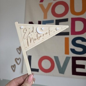 May include: Wooden pennant flag with the words "Eid Mubarak" in script. The flag is decorated with a crescent moon and stars. The flag is held in front of a colorful wall hanging.