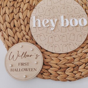 May include: Two wooden signs with a Halloween theme. The larger sign has a white ghost pattern and the text "hey boo" in white letters. The smaller sign has the text "Wilbur's First Halloween" in brown lettering with white stars.
