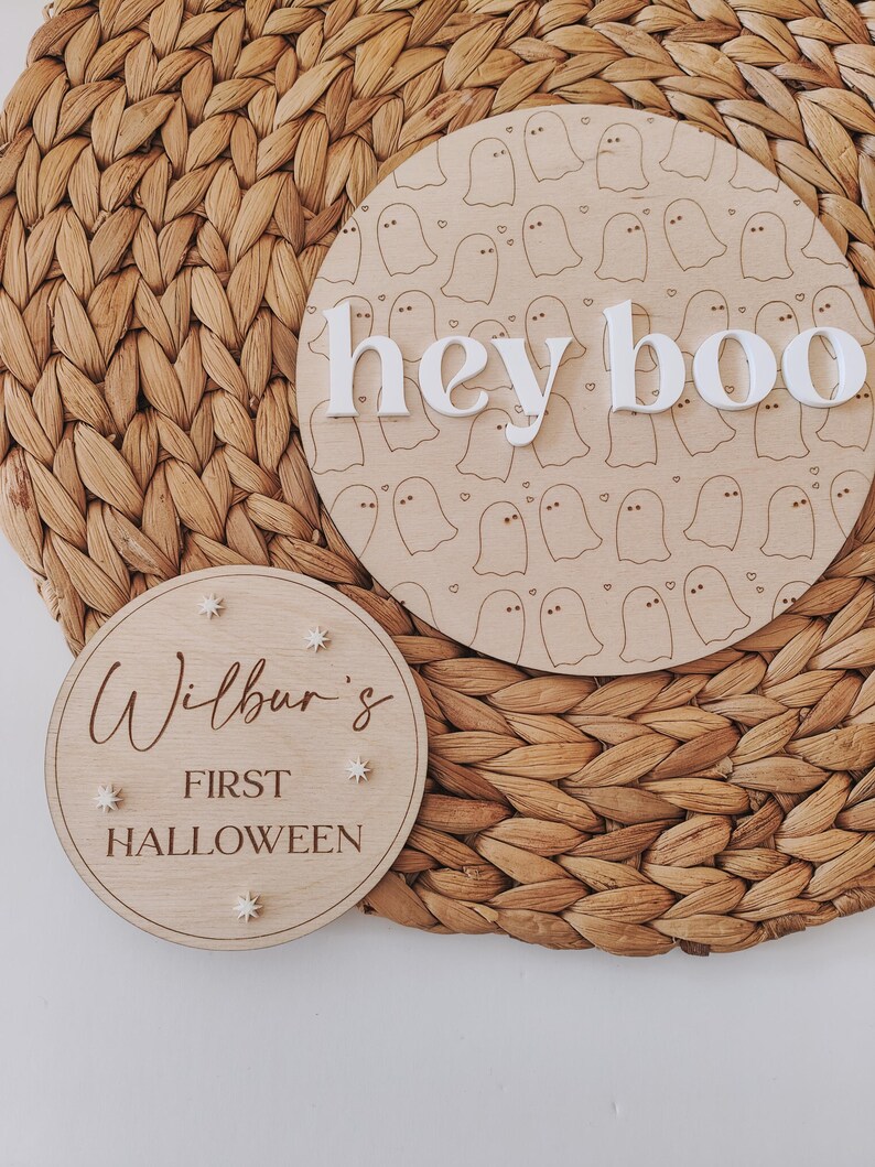 May include: Two wooden signs with a Halloween theme. The larger sign has a white ghost pattern and the text "hey boo" in white letters. The smaller sign has the text "Wilbur's First Halloween" in brown lettering with white stars.
