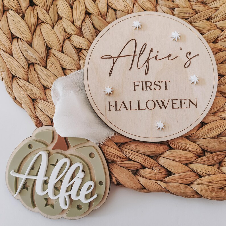 May include: A wooden sign with the text "Alfie's First Halloween" and white stars. A green pumpkin-shaped ornament with the name "Alfie" in white letters.