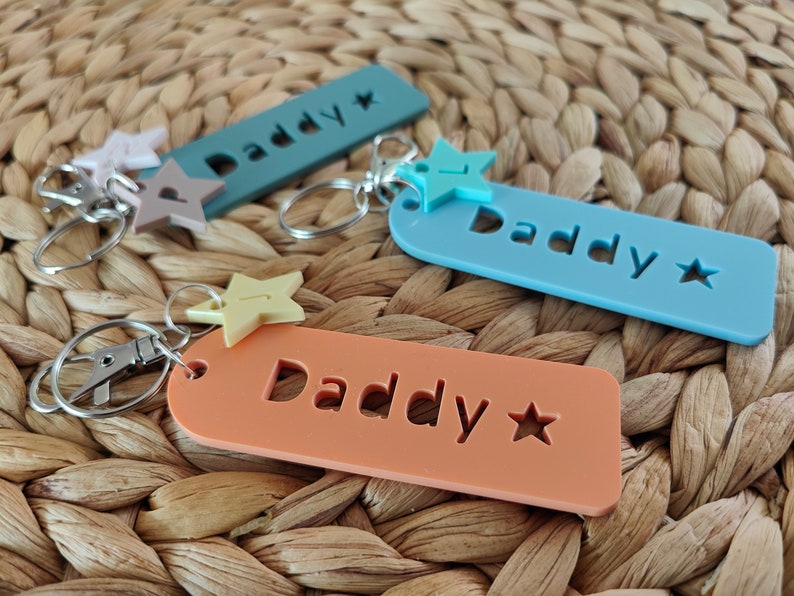 May include: Three keychains with the word "Daddy" cut out of the acrylic. Each keychain has a different color: light blue, blue, and orange. Each keychain has a star charm attached.
