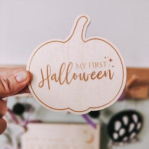 May include: Wooden pumpkin-shaped sign with the text "My First Halloween" in a decorative font.