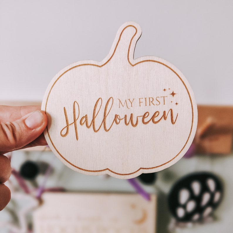 May include: Wooden pumpkin-shaped sign with the text "My First Halloween" in a decorative font.