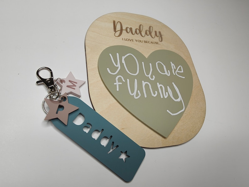 May include: A wooden sign with the text "Daddy I love you because... You are funny" written on it. The sign has a green heart shape attached to it. A blue keychain with the word "Daddy" cut out of it and a star is also included.