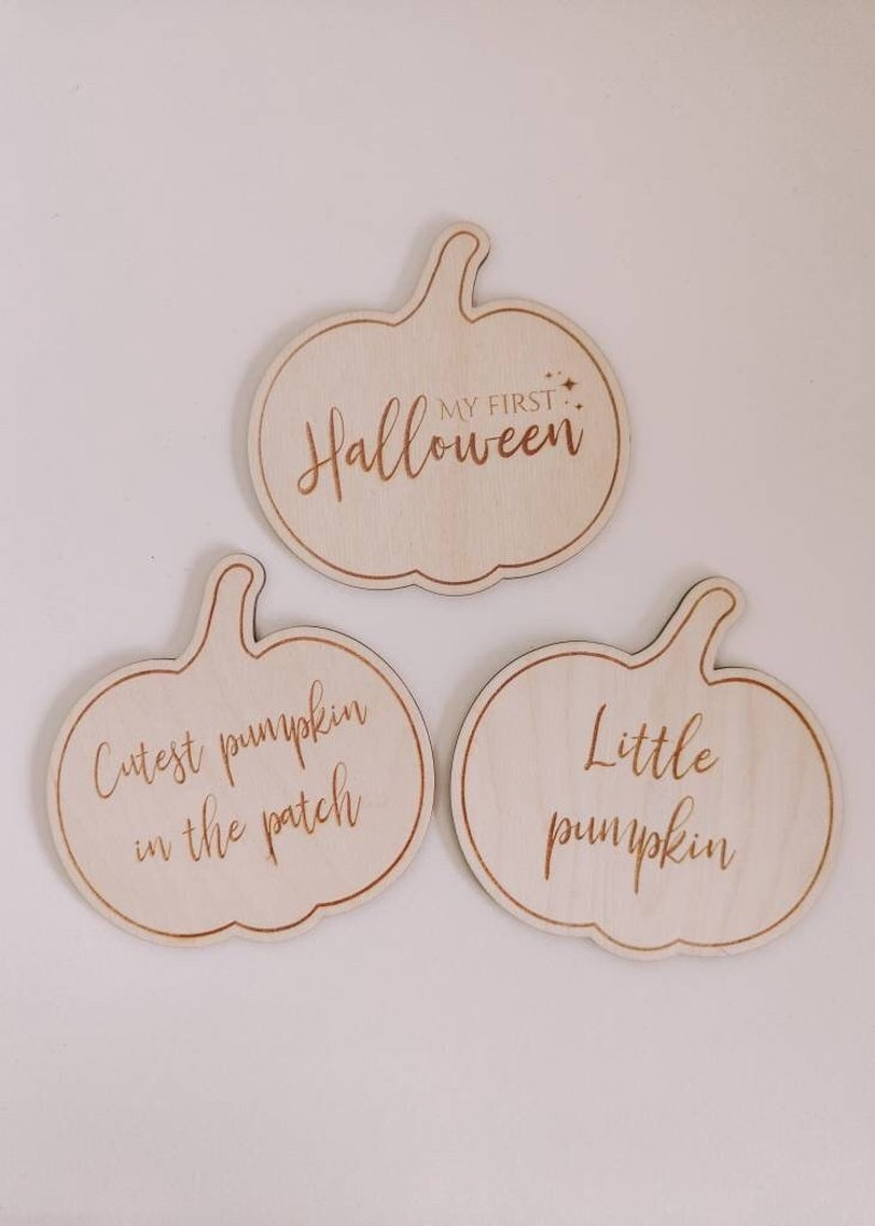 May include: Three wooden pumpkin-shaped signs with different sayings. The top sign says "My First Halloween". The left sign says "Cutest pumpkin in the patch". The right sign says "Little pumpkin".