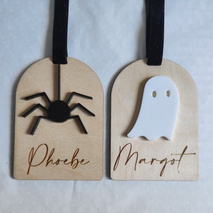May include: Two wooden Halloween ornaments with black ribbon hangers. The ornament on the left has a black spider and the name "Phoebe" written on it. The ornament on the right has a white ghost and the name "Margot" written on it.
