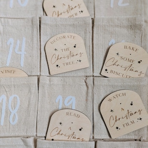 K&ouml;nnte beinhalten: Ein Adventskalender aus Holz mit nummerierten Taschen und Holzetiketten mit weihnachtlichen Aktivit&auml;ten. Die Etiketten haben goldene Schrift und Stern-Akzente. Der Kalender ist aus beigefarbenem Stoff.