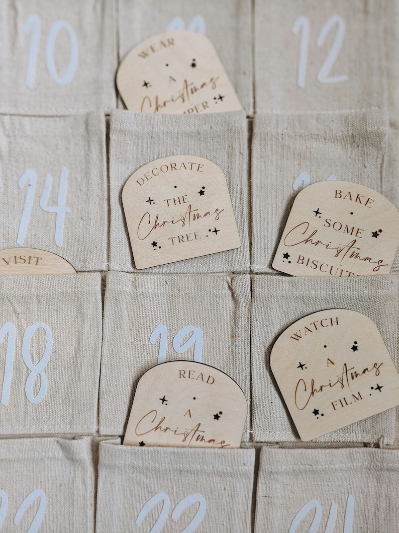 K&ouml;nnte beinhalten: Ein Adventskalender aus Holz mit nummerierten Taschen und Holzetiketten mit weihnachtlichen Aktivit&auml;ten. Die Etiketten haben goldene Schrift und Stern-Akzente. Der Kalender ist aus beigefarbenem Stoff.