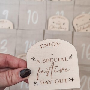 K&ouml;nnte beinhalten: Ein Adventskalender aus Holz mit hellbraunem Hintergrund. Der Kalender hat 24 nummerierte F&auml;cher, jedes mit einem kleinen Holzschild. Das Schild auf dem Bild sagt "Genie&szlig;en Sie einen besonderen festlichen Tag".