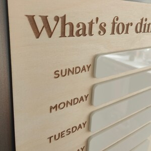 May include: A wooden sign with the text "What's for dinner?" and blank white spaces for each day of the week: Sunday, Monday, Tuesday, Wednesday, Thursday, Friday, and Saturday.