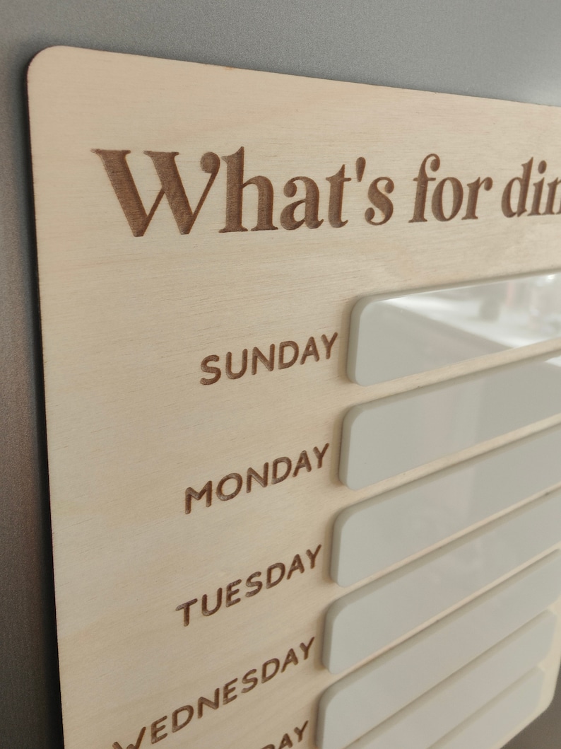 May include: A wooden sign with the text "What's for dinner?" and blank white spaces for each day of the week: Sunday, Monday, Tuesday, Wednesday, Thursday, Friday, and Saturday.