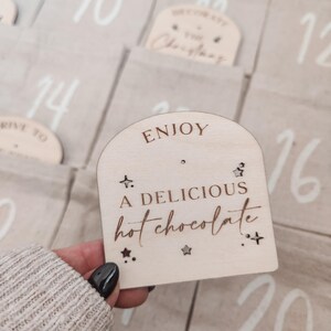 K&ouml;nnte beinhalten: Eine Holzt&uuml;r f&uuml;r einen Adventskalender mit dem Text "ENJOY A DELICIOUS hot chocolate" und Sternen. Die T&uuml;r ist hellbraun mit wei&szlig;em Text und Sternen.