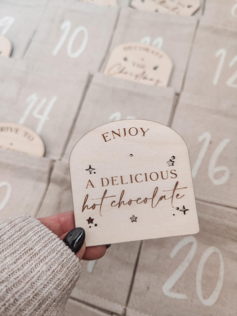 K&ouml;nnte beinhalten: Eine Holzt&uuml;r f&uuml;r einen Adventskalender mit dem Text "ENJOY A DELICIOUS hot chocolate" und Sternen. Die T&uuml;r ist hellbraun mit wei&szlig;em Text und Sternen.