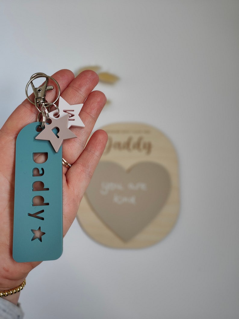 May include: A teal blue keychain with the word "Daddy" and a star cutout. The keychain has two small stars attached to it, one white and one pink.