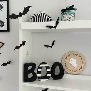 May include: Black and white striped BOO letters with a black and white striped pumpkin, a black and white ghost sign that says "stay spooky stay spooky stay spooky stay", and black bats on a white shelf.