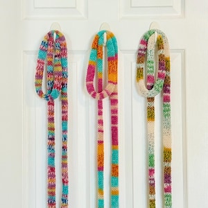 May include: Three long, thin, hand-knitted scarves hanging vertically. The first scarf features a vibrant mix of turquoise, pink, yellow, and purple. The second scarf displays stripes of pink, orange, and turquoise. The third scarf has a cream base with green, pink, and yellow accents.