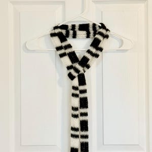 May include: A long, narrow knit scarf featuring a repeating pattern of bold black and white horizontal stripes. The scarf is constructed with a visible yarn texture and is tied in a simple knot at the centre, hanging down in two equal lengths.