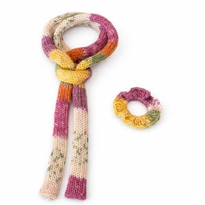 Puede incluir: Una colorida bufanda de punto y una scrunchie a juego. La bufanda está anudada y presenta rayas rosas, naranjas, amarillas, verdes y crema. La scrunchie tiene una paleta de colores similar. Ambos artículos están hechos de un hilo suave y texturizado.