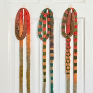 May include: Three long, thin, knitted scarves hanging vertically. The first is an orange and yellow gradient. The second features horizontal stripes of green, orange, and brown. The third has a textured chain-link pattern in green and orange.