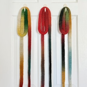 May include: Three long, thin, knitted scarves hanging vertically. The first scarf features a gradient of cream, green, yellow, and red. The second scarf displays a gradient of red, brown, and dark green. The third scarf shows a gradient of dark green, red, yellow, cream, and light blue.