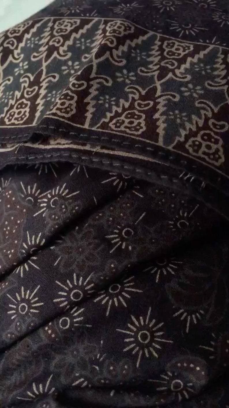 Puede incluir: Una tela marr&oacute;n oscura con un patr&oacute;n blanco de peque&ntilde;as estrellas y formas m&aacute;s grandes que se asemejan al sol. La tela tiene una superficie texturizada, ligeramente rugosa.