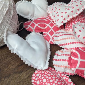May include: A collection of handmade fabric hearts in various pink and white patterns. The hearts are sewn with a white lace trim and are arranged on a wooden surface.