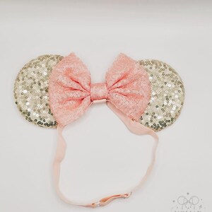 May include: A headband featuring two circular ears covered in shimmering gold sequins and a large, central bow made of light pink sequins. The headband includes an adjustable light pink elastic strap with a metal slider for a secure fit.