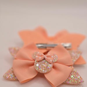 May include: A peach-colored hair bow featuring a layered design with glittery accents and a central Minnie Mouse-shaped embellishment with white polka dots. The accessory includes a metal alligator clip on the back and a small Nomad Mouse logo.