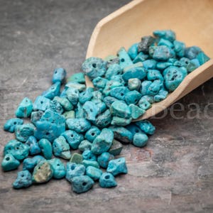 May include: A wooden scoop overflowing with turquoise-colored, irregular-shaped beads. The beads are scattered on a textured, gray surface. The image includes the text "Kira Beads".