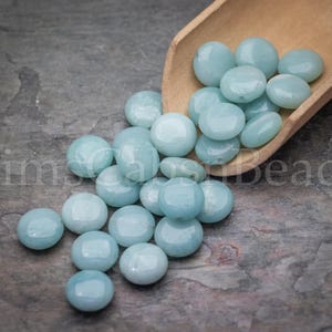 May include: A collection of light blue, round beads, some spilling from a small wooden scoop. The beads have a smooth, glossy finish and are scattered on a textured, gray surface. The image includes the text "Kim's Beads".