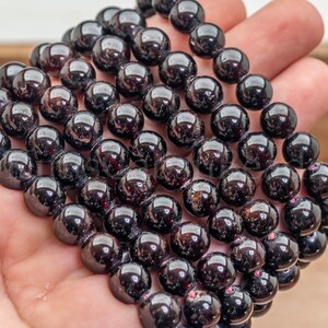 May include: A close-up of a strand of polished, dark red garnet beads. The beads are round and have a glossy finish, reflecting light. The beads are held in a hand, showcasing their color and texture. The beads are approximately 8mm in diameter.