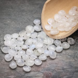 May include: A collection of faceted, translucent moonstone beads scattered on a dark gray surface. A wooden scoop holds additional beads. The beads have a pearlescent sheen and a variety of shapes, perfect for jewelry making.