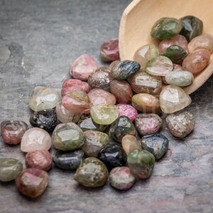 May include: A collection of tumbled gemstones in various colors, including pink, green, brown, and clear. The gemstones are spilling from a small wooden scoop onto a gray surface. The stones have a smooth, polished appearance.