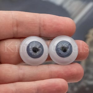 May include: Two glass doll eyes with a light blue iris and black pupil. The eyes are round and have a white sclera. The eyes are held in a person's hand.