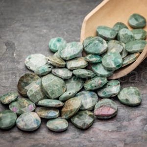May include: A collection of green and pink gemstone beads, with a faceted cut, spilling from a small wooden scoop onto a dark gray surface. The beads vary in size and shade, with some showing hints of pink.