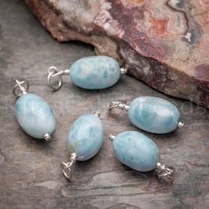 May include: Five oval-shaped, light blue larimar gemstone pendants with silver-colored findings. The gemstones have a marbled appearance with white and blue swirls. The pendants are displayed on a textured, gray and brown surface.