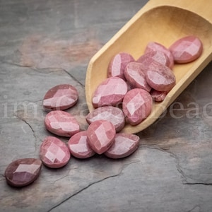 May include: A wooden scoop filled with pink, faceted, teardrop-shaped gemstone beads. The beads are scattered on a gray, textured surface.